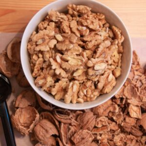 Top view of a bowl filled with shelled walnuts surrounded by walnut shells on a wooden table.