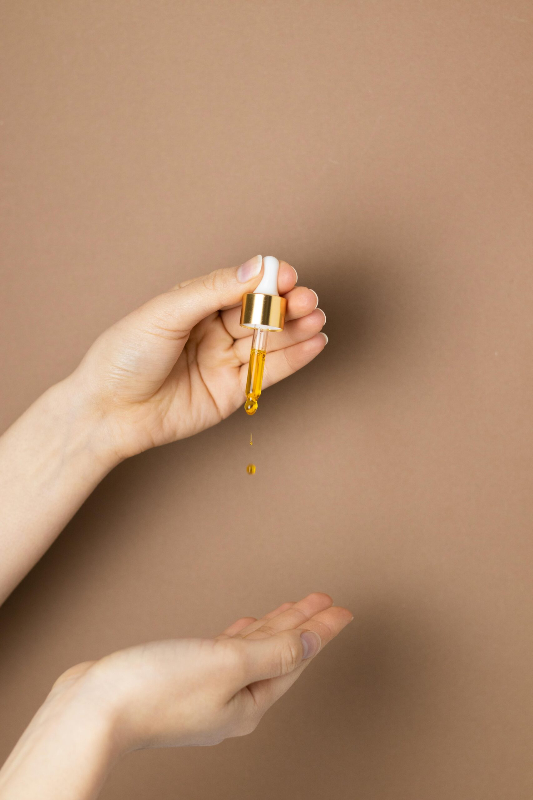 Close-up of hands applying skincare serum with a dropper on brown background.